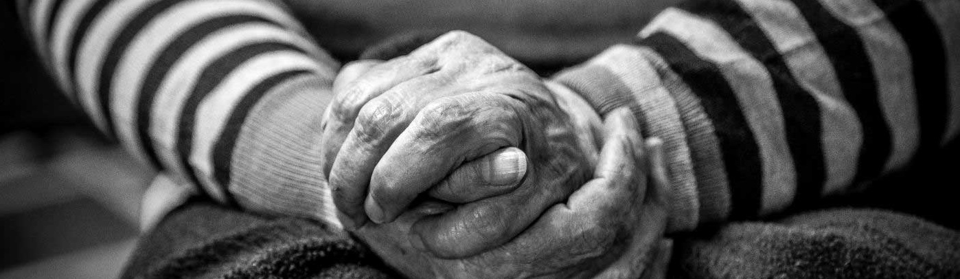 Close-up black and white image of elderly hands clasped together. The person wears a striped sweater, conveying warmth and reflection.