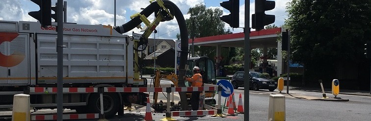 Construction workers operate heavy machinery with a vacuum truck at a road intersection. Safety barriers and cones are set up under a partly cloudy sky.
