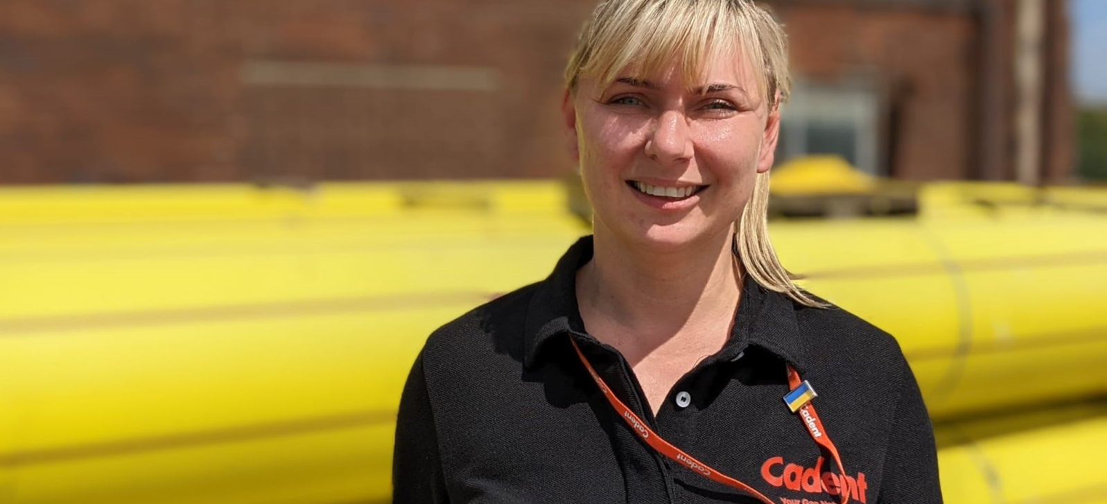 Smiling woman in a black Cadent shirt stands outdoors in front of stacked yellow pipes. The setting is industrial with bright natural light.
