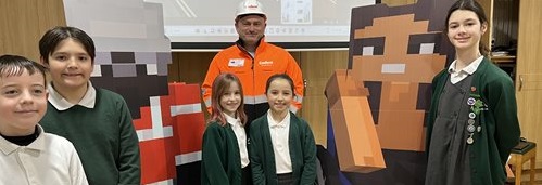 A group of five children in school uniforms stands with a man in an orange safety jacket and helmet. They pose next to two large Minecraft character cutouts, smiling. A screen in the background shows a pixelated game scene, creating a cheerful and educational atmosphere.