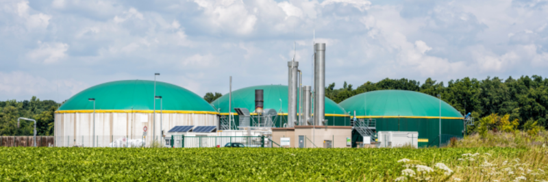 A rural biomethane plant with green-domed digesters and metal chimneys, surrounded by crop fields and trees, under a partly cloudy sky, showcasing sustainable energy production.