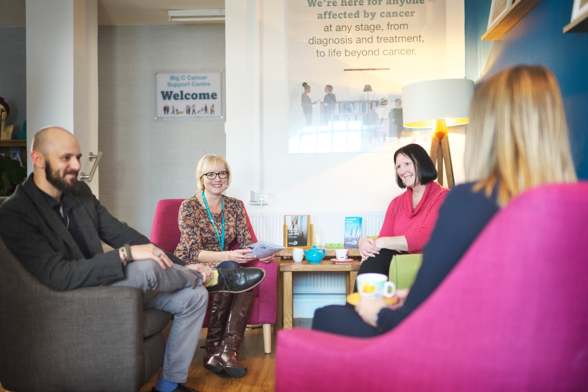 Four people sit in a welcoming support center, engaging in friendly conversation, surrounded by colorful chairs and informative posters about cancer support.