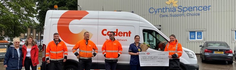 A group of people stand cheerfully in front of a white van with 'Cadent' branding. They hold a large check and a bouquet, near a building marked 'Cynthia Spencer.' Rainy weather visible on the ground.