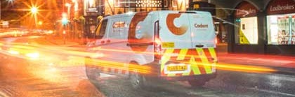 A white van with red and yellow stripes and a logo displaying "Cadent" is on a wet street at night, with surrounding light trails from traffic.