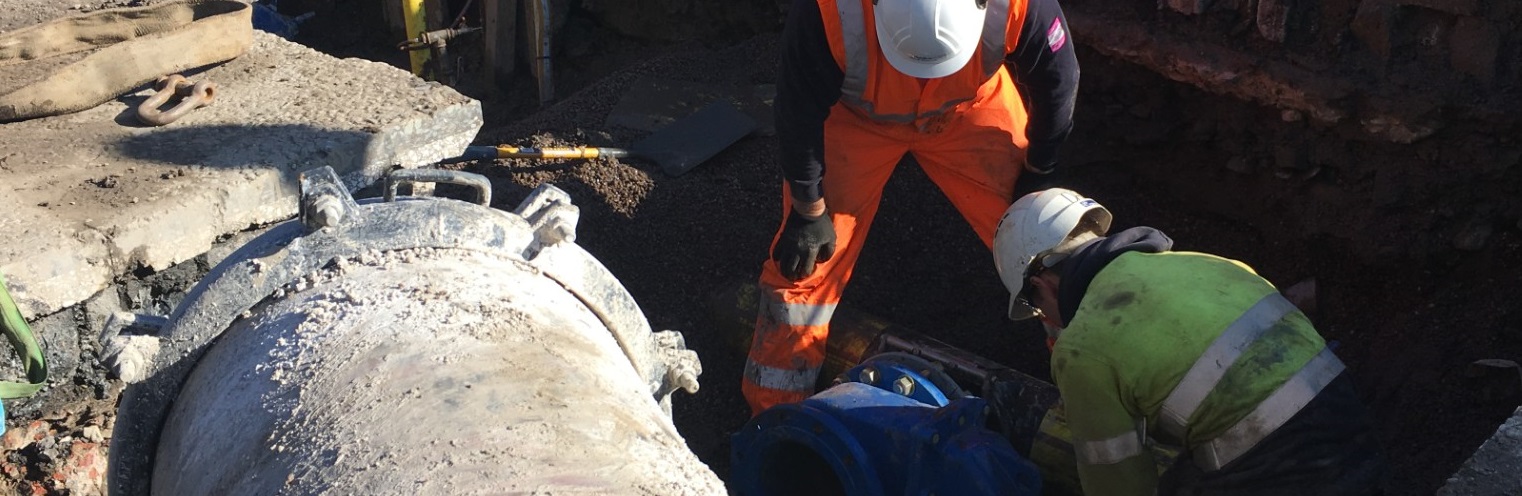Two construction workers in high-visibility jackets and helmets work on a large underground pipe in a sunlit trench, focusing intently on their task_banner_Image