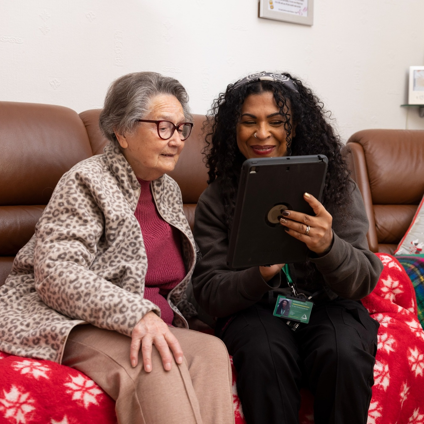 Elderly woman in a leopard print jacket and younger woman with curly hair sit on a sofa, smiling and looking at a tablet together, conveying warmth.