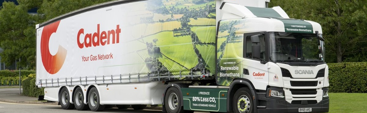 A large white truck with a trailer featuring the Cadent logo and the text 'Your Gas Network.' It is parked on a road, surrounded by green trees. The truck advertises 'Power by Renewable Biomethane' and '80% Less CO2.' The image conveys an eco-friendly transport theme.