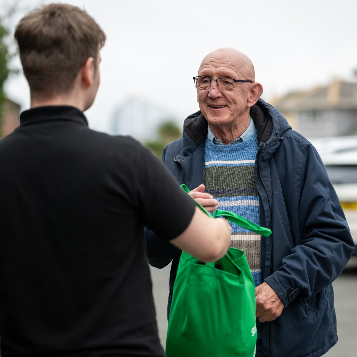A smiling older man in glasses and a blue jacket receives a green bag from a younger person outdoors. The scene conveys warmth and community.