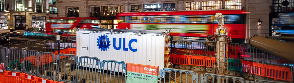 Nighttime city street with construction barriers and a white container labeled ULC, surrounded by orange fencing, with red double-decker buses passing in front of a large illuminated building.
