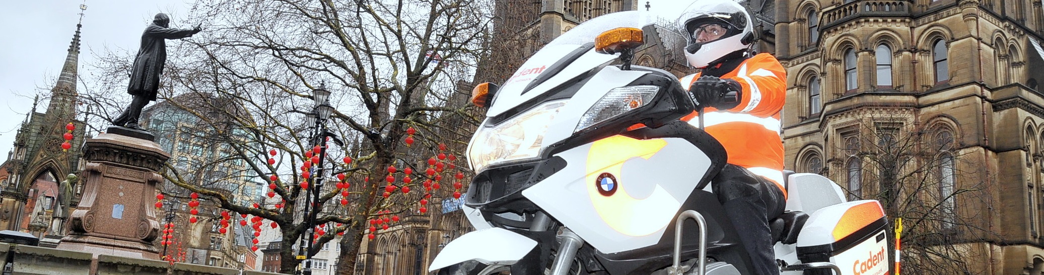 Motorcyclist in orange gear rides near a historic building and statue, with red lanterns hanging from trees, creating a dynamic urban scene.