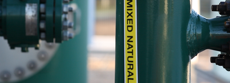Close-up of a green industrial pipe with a yellow label reading 'Mixed Natural Gas.' A bolted flange is visible in the blurred background.