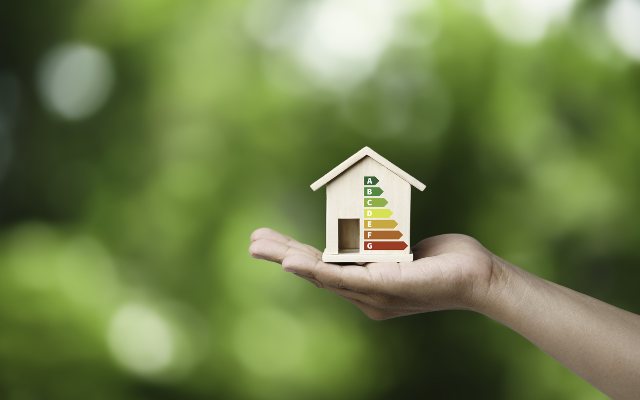 Small wooden house with an A-G energy efficiency rating displayed on the front, resting on an open hand against a green blurred background.
