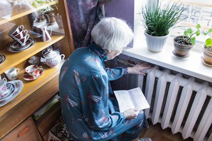 An elderly person in a blue patterned robe sits by a window, checking a radiator. Nearby, potted plants and a cabinet of teacups create a cozy atmosphere.