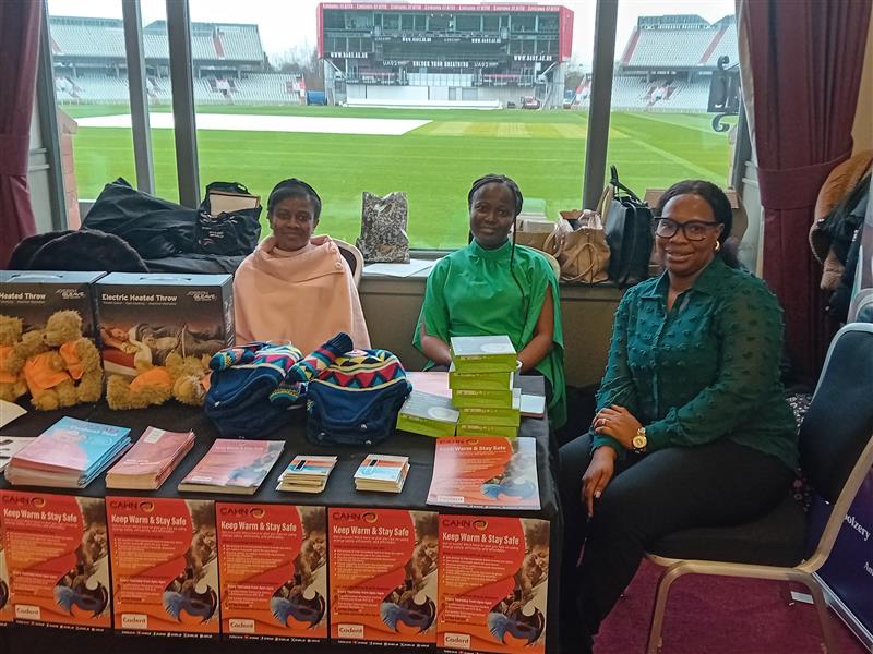 Three women sit behind a table covered with brochures, colorful blankets, and gift boxes in a stadium lounge. The setting is warm and inviting_image