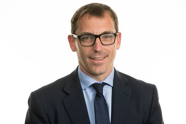 A man in a suit and tie smiles warmly against a plain white background. He wears glasses and exudes a professional and approachable demeanor.