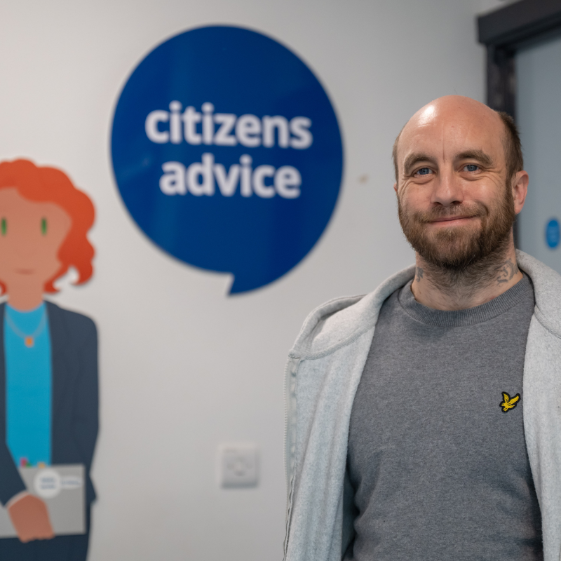 A person stands indoors in front of a wall with a large Citizens Advice speech‑bubble logo and an illustrated figure holding a folder. The person is wearing a grey top and a light-coloured hoodie. The setting appears to be a Citizens Advice office, with a doorway and noticeboard visible in the background.