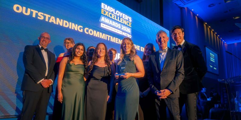 A smiling group in formalwear poses on a blue-lit stage, holding an award beneath a projection reading “Employer Excellence Awards 2026.”