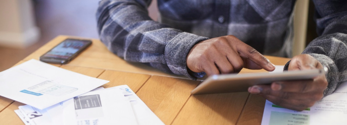Person sitting at a wooden table using a tablet, with papers and a smartphone nearby