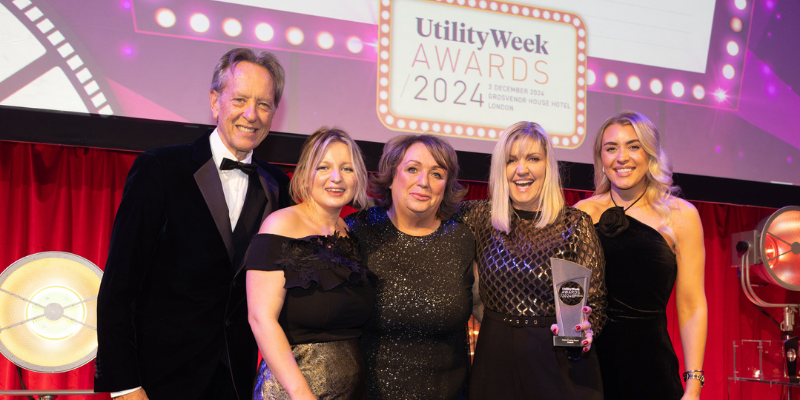 A group of people posing with a glass trophy on hand with Utility Week Awards board on the background.