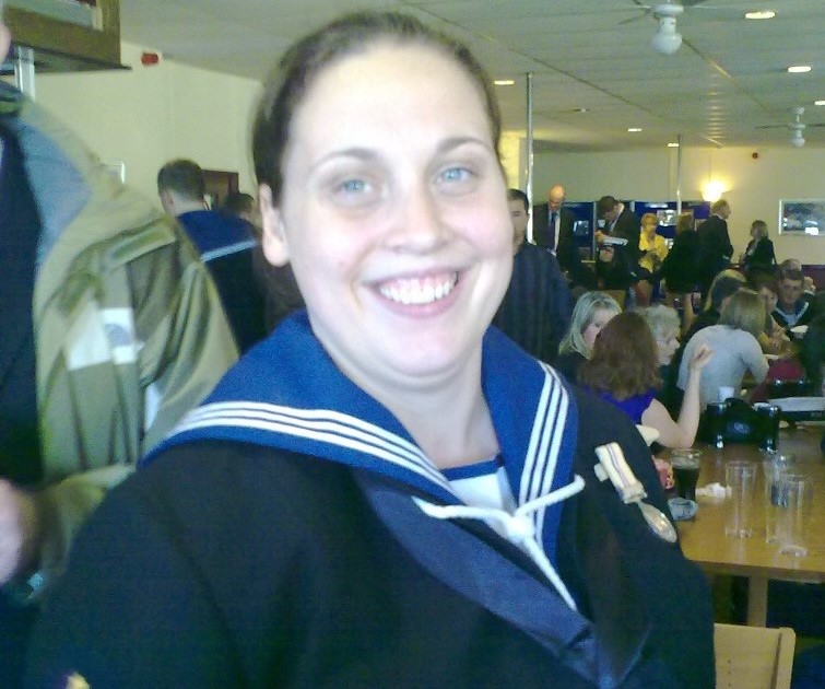 Smiling person in a navy uniform with medals in a crowded room, suggesting a celebratory event. The atmosphere is lively and social.