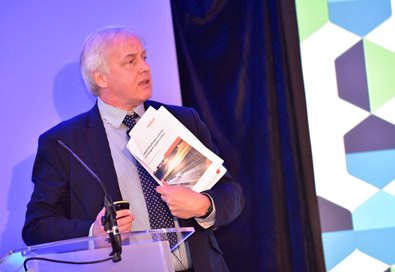A man in a suit speaks at a podium, holding documents. The background features geometric patterns in blue and green, suggesting a professional event.