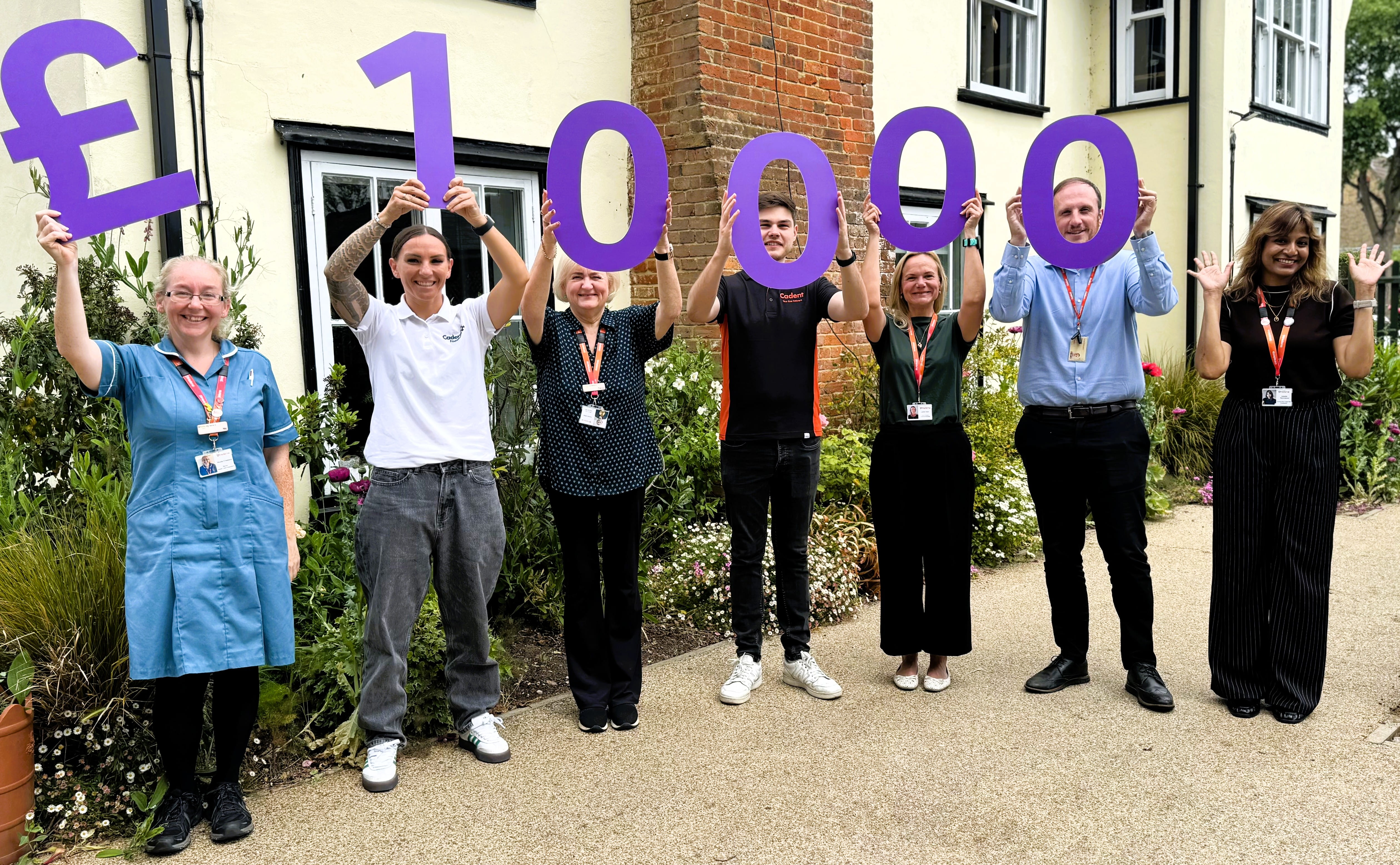 Seven people stand outside a building holding large purple characters displaying £10000. They are smiling, conveying celebration and achievement.