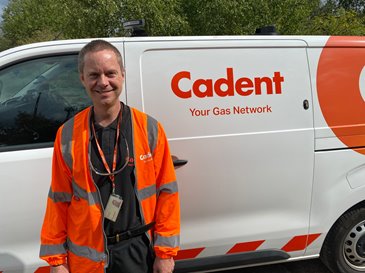 Man in an orange high-visibility jacket stands smiling in front of a white utility van with 