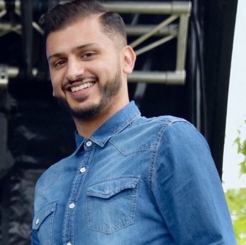 Gurpreet - Smiling person in a blue denim shirt, stands in an outdoor setting with blurred dark structures in the background, exuding a cheerful vibe.