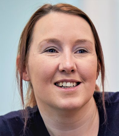 Smiling woman with brown hair in a blue shirt, gazing slightly to the left. The background is softly blurred, creating a calm and friendly atmosphere.
