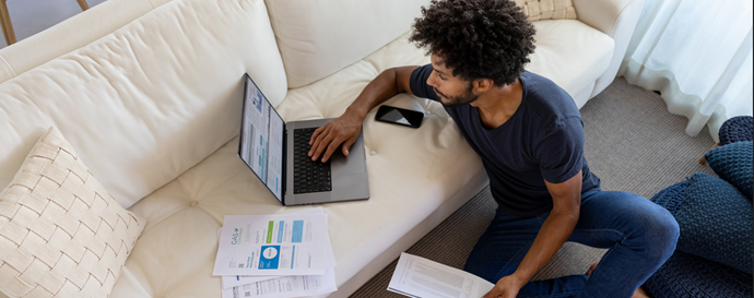 Person sitting on the floor beside a sofa, reviewing printed reports while using a laptop on the couch. A smartphone and additional documents are nearby in a bright living room setting.