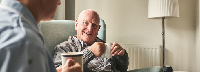 Smiling senior man with cup of coffee talking to female caregiver sitting on the sofa