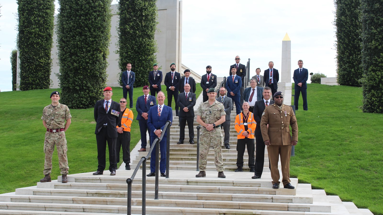 A group of people, including military personnel in camouflage and uniforms, stand solemnly on steps in front of tall trees and a memorial, conveying respect.