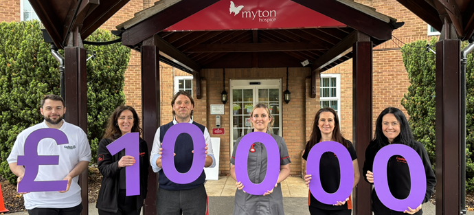 Six people smiling, holding large purple numbers reading £100,000 in front of a building with a sign myton hospice. They appear joyful and proud.