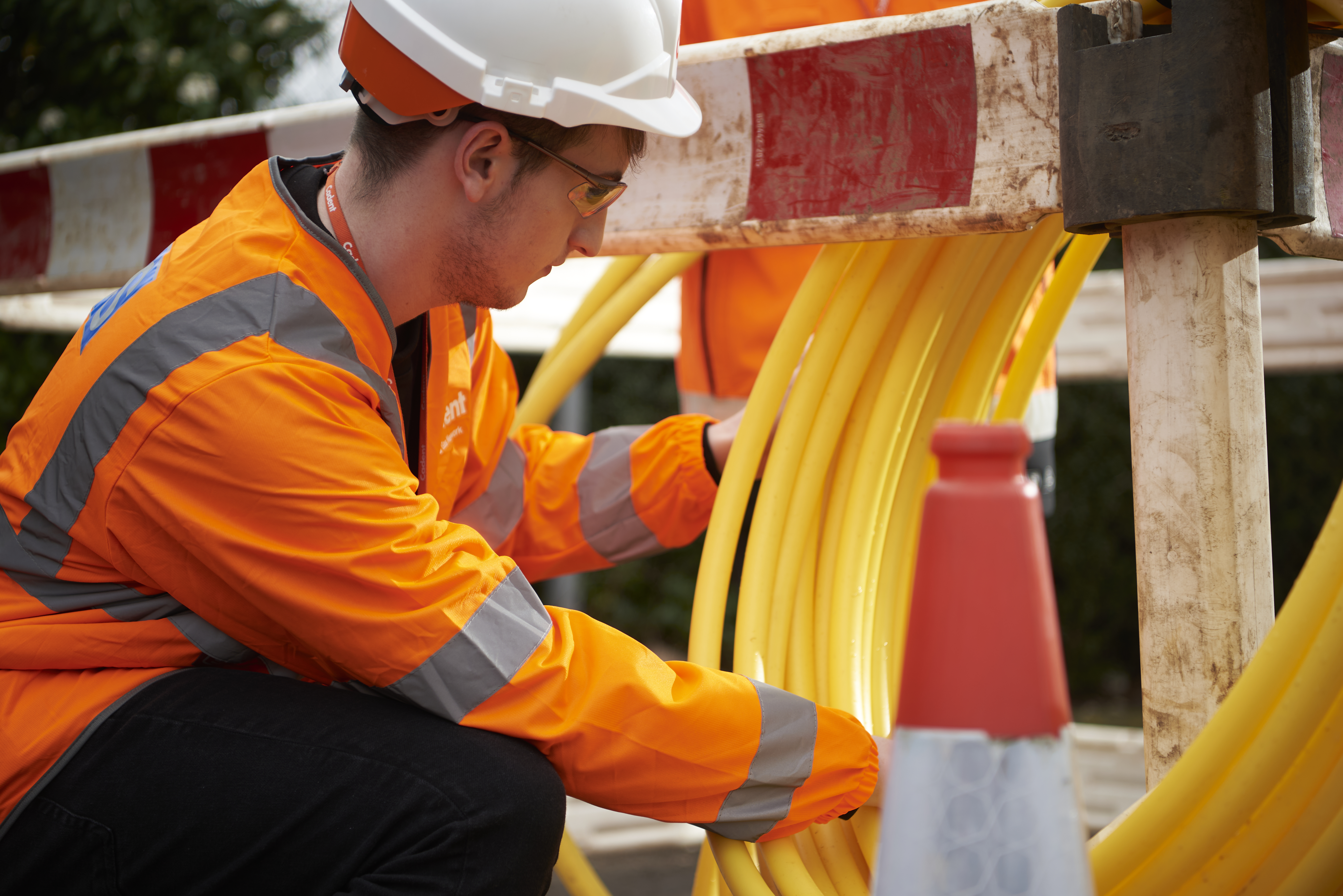 Construction worker in orange high-visibility gear and hard hat installs bright yellow cables behind a red-and-white road barrier.