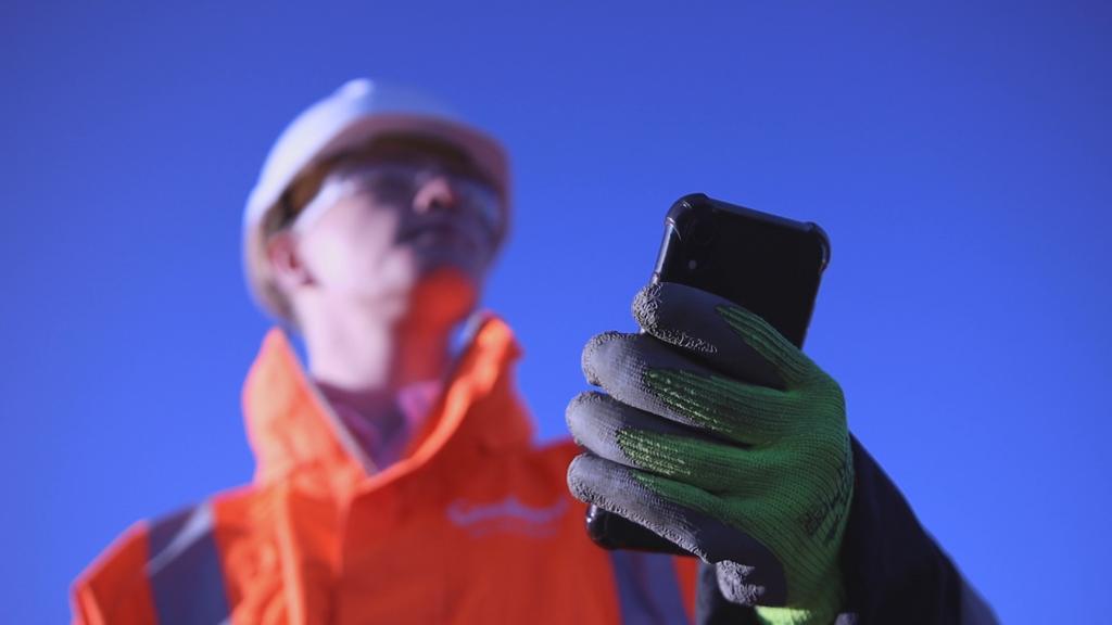 A construction worker in an orange safety jacket and white helmet looks up at a smartphone. Gloved hand holds the phone against a clear blue sky background.