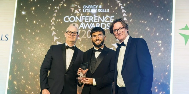 Three people standing and posing for a picture with a trophy on hand at the centre on stage of an Award show.