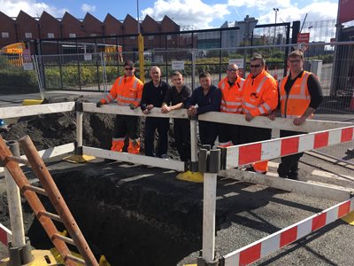 A group of seven people in safety gear stand behind a barricade surrounding a large construction pit on a sunny day, conveying teamwork and focus.