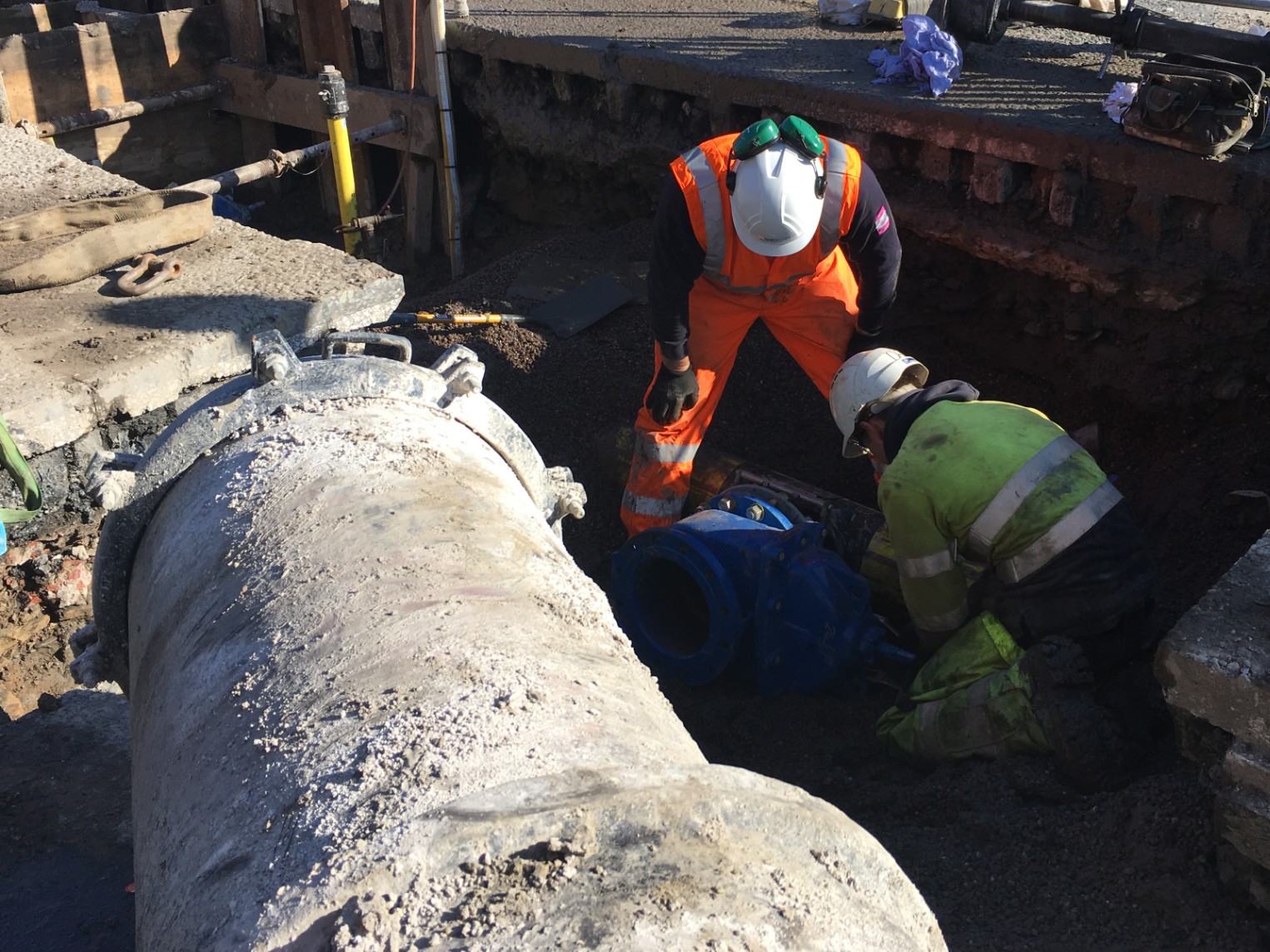 Two construction workers in high-visibility jackets and helmets work on a large underground pipe in a sunlit trench, focusing intently on their task_image