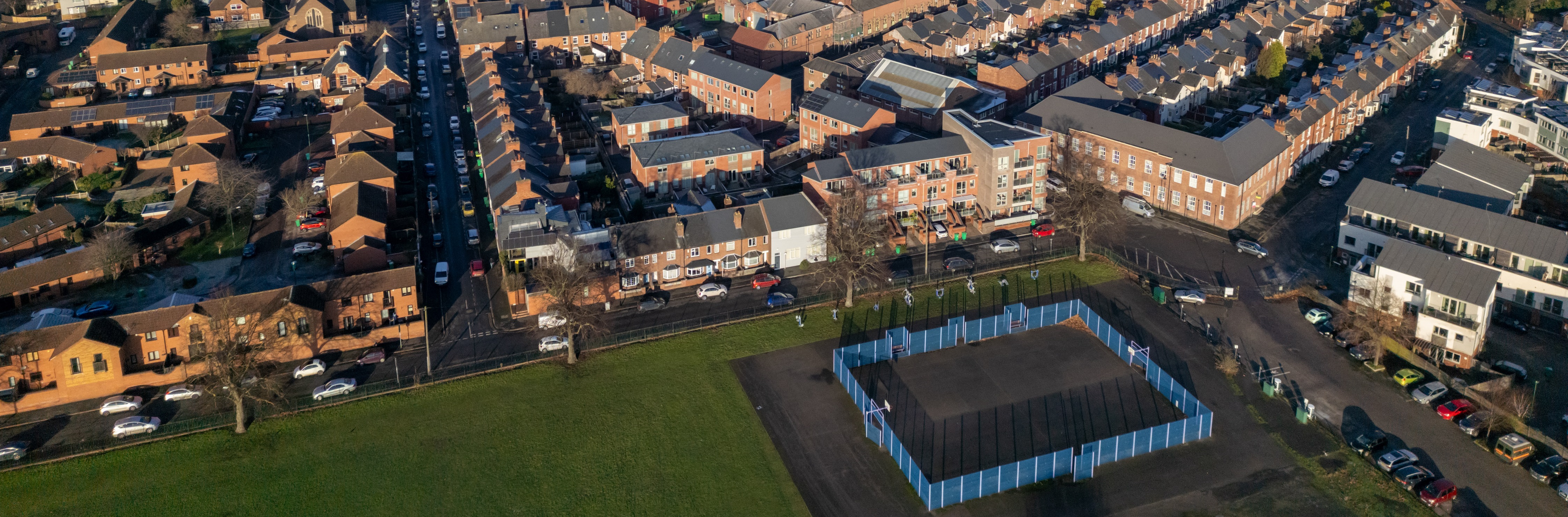 Aerial view of a cityscape with rows of residential houses and streets. A fenced sports court is in the foreground, and industrial buildings are in the distance. The atmosphere is calm and clear.