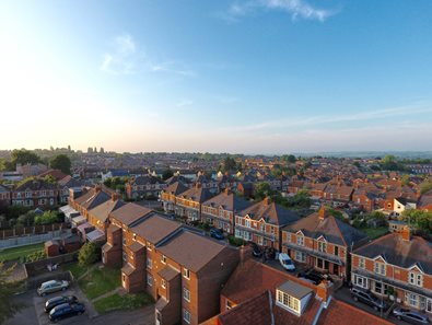 Aerial view of a residential neighborhood with rows of brick houses, tiled roofs, and parked cars under a clear blue sky