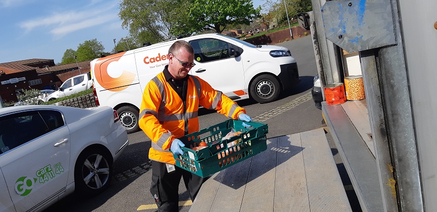 Person in a bright orange safety jacket and gloves loading a green crate of food items onto a truck, with Cadent and Cadent vehicles parked nearby.