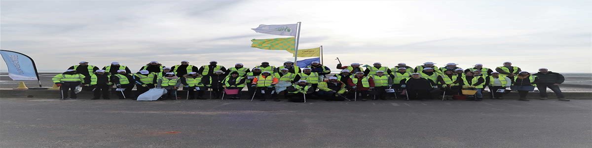 A group of people wearing high-visibility vests, holding litter pickers, pose outdoors under cloudy skies. Several flags are raised behind them.