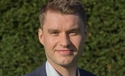 A person wearing a suit smiles warmly at the camera, standing outdoors in front of a lush green hedge, conveying a professional and approachable tone.