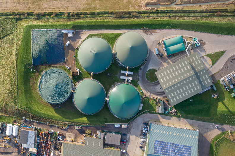 Aerial view of a biogas facility with five large, green, dome-shaped tanks, surrounded by buildings and pathways, set in a grassy landscape.