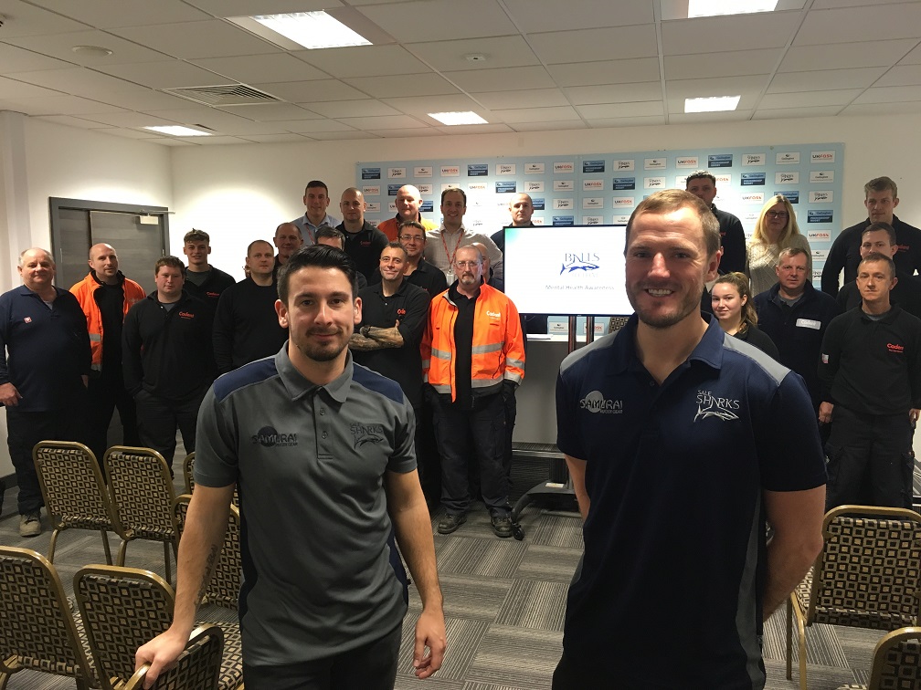 Two men in branded shirts stand in front of a group of people in a conference room. A screen displays a logo. The mood is professional and upbeat.