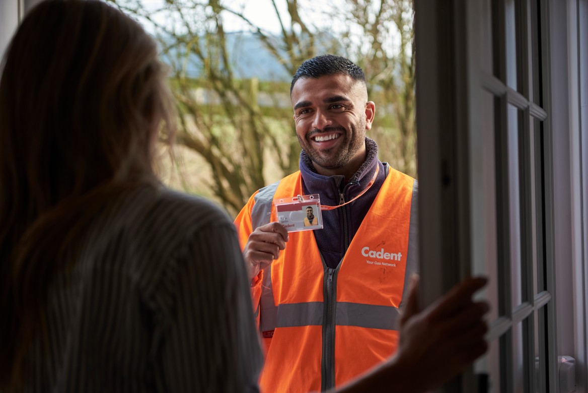 A man in an orange safety vest smiles while holding an ID badge and stands outside a doorway, speaking to a person inside. The mood is friendly and professional.