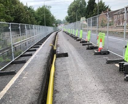 A paved road under construction is fenced off on both sides. A yellow pipe runs through a trench in the road, with safety signs and cones nearby.