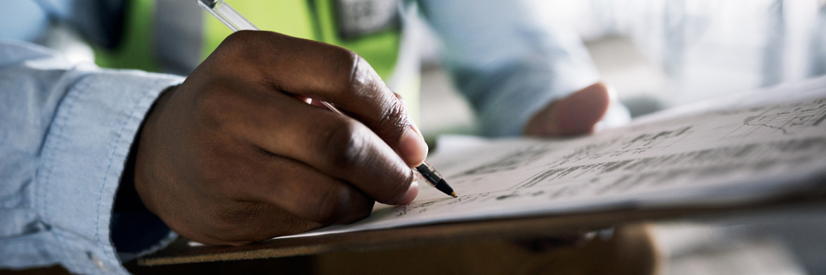 A person writing on a sheet of paper clipped to a wooden clipboard. They hold a pen in their right hand. The background is blurred, and the individual is wearing a light blue shirt and a reflective vest.