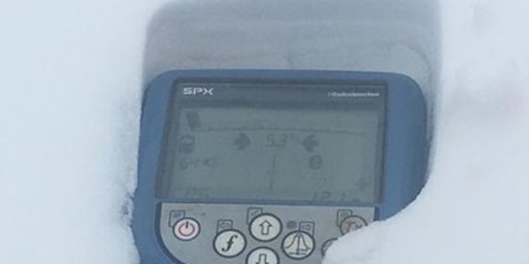 A digital device with a blue frame is partially buried in snow. The screen displays measurements, and it conveys resilience in harsh, snowy conditions.