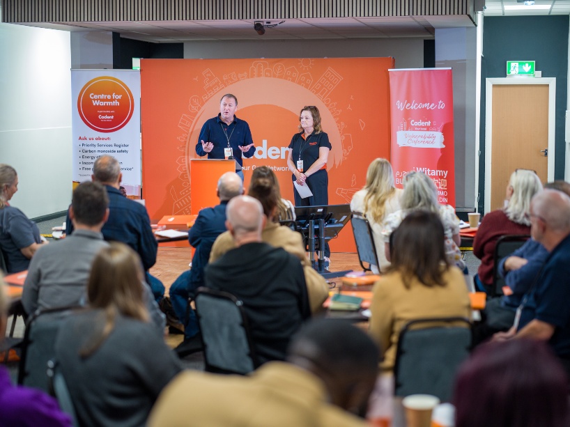 Two presenters stand on a stage in front of an orange backdrop titled 